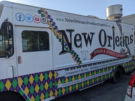 A New Orleans-themed food truck is parked on a street. It features vibrant designs, including Mardi Gras bead graphics and a fleur-de-lis motif. The truck's side promotes corporate catering, weddings, festivals, and other events, with contact information for locations like Harvey and Covington. Social media icons are displayed at the top left corner.