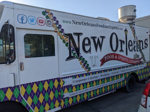 A New Orleans-themed food truck is parked on a street. It features vibrant designs, including Mardi Gras bead graphics and a fleur-de-lis motif. The truck's side promotes corporate catering, weddings, festivals, and other events, with contact information for locations like Harvey and Covington. Social media icons are displayed at the top left corner.