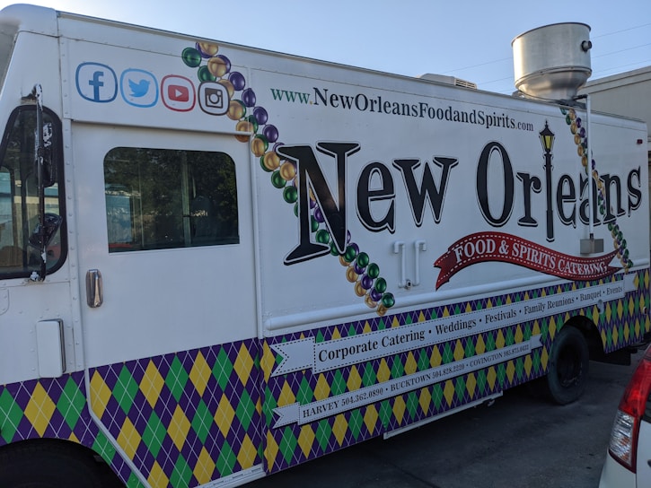 A New Orleans-themed food truck is parked on a street. It features vibrant designs, including Mardi Gras bead graphics and a fleur-de-lis motif. The truck's side promotes corporate catering, weddings, festivals, and other events, with contact information for locations like Harvey and Covington. Social media icons are displayed at the top left corner.