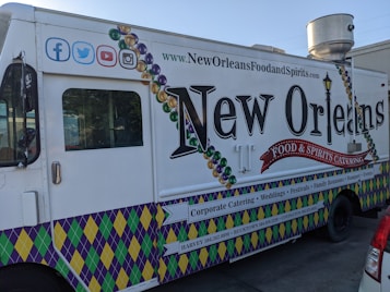 A New Orleans-themed food truck is parked on a street. It features vibrant designs, including Mardi Gras bead graphics and a fleur-de-lis motif. The truck's side promotes corporate catering, weddings, festivals, and other events, with contact information for locations like Harvey and Covington. Social media icons are displayed at the top left corner.