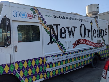 A New Orleans-themed food truck is parked on a street. It features vibrant designs, including Mardi Gras bead graphics and a fleur-de-lis motif. The truck's side promotes corporate catering, weddings, festivals, and other events, with contact information for locations like Harvey and Covington. Social media icons are displayed at the top left corner.