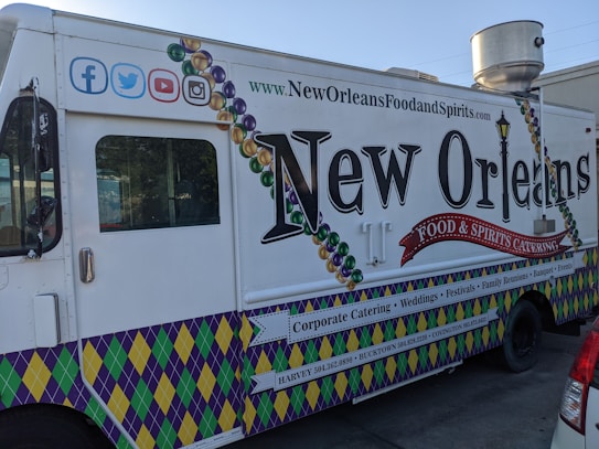 A New Orleans-themed food truck is parked on a street. It features vibrant designs, including Mardi Gras bead graphics and a fleur-de-lis motif. The truck's side promotes corporate catering, weddings, festivals, and other events, with contact information for locations like Harvey and Covington. Social media icons are displayed at the top left corner.