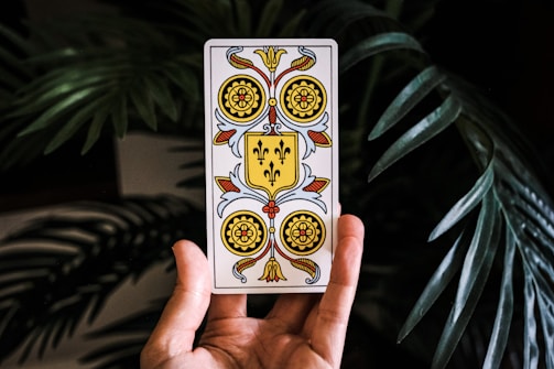 A hand is holding a tarot card with ornate designs featuring a crest in the center. The card displays a symmetrical pattern with floral motifs and a prominent shield-shaped emblem adorned with fleur-de-lis. The background includes large green leaves, possibly from a tropical plant, adding a natural setting to the foreground.