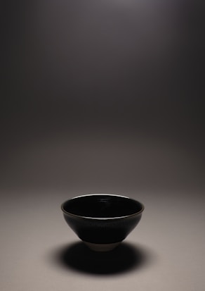 A single, elegant ceramic bowl showcased under dim lighting, casting a soft shadow on a neutral surface. The bowl appears to have a glossy finish, reflecting the subtle light above.
