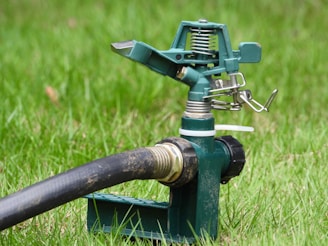 A green metal garden sprinkler is positioned on the grass, connected to a black hose. The sprinkler has intricate metal parts including a spring and levers, designed to distribute water across a lawn.