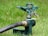 A green metal garden sprinkler is positioned on the grass, connected to a black hose. The sprinkler has intricate metal parts including a spring and levers, designed to distribute water across a lawn.