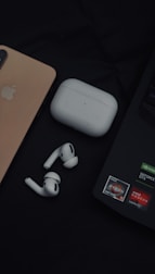 A close-up of various electronic gadgets on a dark surface, including a gold smartphone, a pair of wireless earbuds, their white charging case, and a laptop with several stickers.