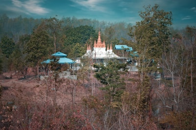 A secluded structure with blue roofs and a central spire is surrounded by dense trees and a dry landscape. The scene has a serene and tranquil atmosphere, with soft light filtering through the foliage.