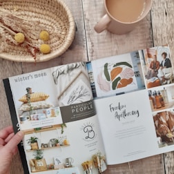 A hand holds open a lifestyle magazine on a rustic wooden table. The left page features interior design elements, including cushions and plant decor, along with contact information. The right page showcases artwork and a feature on Freedom Apothecary, highlighting two seated individuals and a collection of amber bottles. A woven basket with dried flowers and a mug filled with tea or coffee are placed nearby, complementing the cozy and rustic atmosphere.