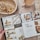 A hand holds open a lifestyle magazine on a rustic wooden table. The left page features interior design elements, including cushions and plant decor, along with contact information. The right page showcases artwork and a feature on Freedom Apothecary, highlighting two seated individuals and a collection of amber bottles. A woven basket with dried flowers and a mug filled with tea or coffee are placed nearby, complementing the cozy and rustic atmosphere.