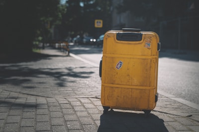 A large, worn yellow suitcase with a handle and wheels is positioned on a paved sidewalk. The suitcase features a sticker with '100%' on it and there are shadows of trees cast on the ground around it. In the background, a street is visible with blurred vehicles and road signs, conveying a sense of an urban setting.