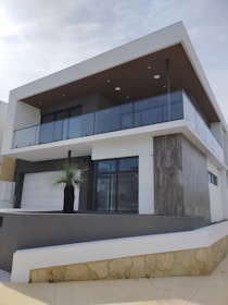 A modern, two-story building with a minimalist design features a flat roof and large glass windows. The upper level includes a balcony enclosed with transparent glass railings. The façade incorporates a combination of white and gray tones with a touch of natural wood and stone elements.
