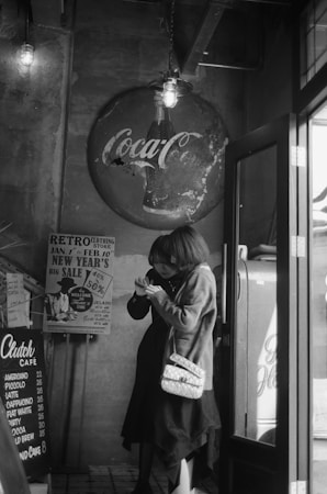 A dimly lit retro-style cafe interior features vintage decor including a weathered Coca-Cola sign and a menu with coffee options. Two women are engaged with a smartphone, one carrying a white quilted bag. Nostalgic advertisement posters are visible in the background.