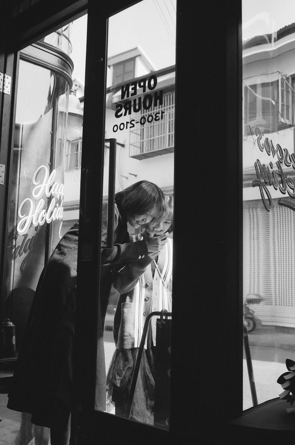 grayscale photo of woman in black coat standing beside glass window