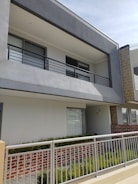 A modern two-story building facade with a minimalist design, featuring a prominent balcony with metal railings. The exterior consists of gray and white walls, with some sections of exposed red brick. There is a neatly maintained garden with green plants along the base.