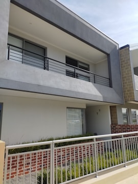A modern two-story building facade with a minimalist design, featuring a prominent balcony with metal railings. The exterior consists of gray and white walls, with some sections of exposed red brick. There is a neatly maintained garden with green plants along the base.