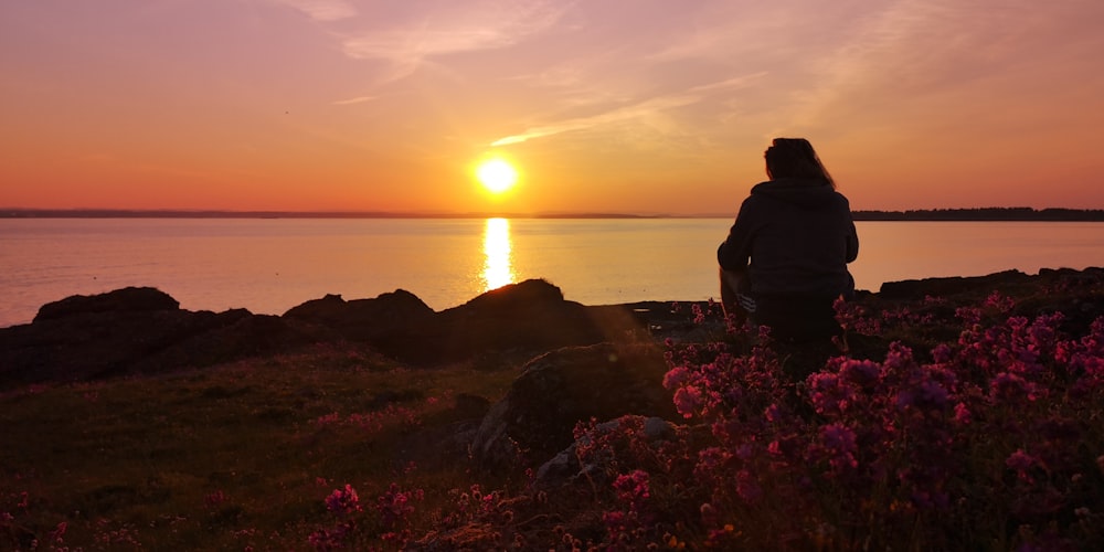 silhouette of person sitting on rock near body of water during sunset