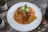 A plate of spaghetti topped with a rich tomato and meat sauce, garnished with grated cheese and fresh basil leaves. The dish is presented on a white plate placed on a rustic wooden table, surrounded by small potted plants.