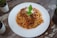 A plate of spaghetti topped with a rich tomato and meat sauce, garnished with grated cheese and fresh basil leaves. The dish is presented on a white plate placed on a rustic wooden table, surrounded by small potted plants.