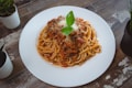 A plate of spaghetti topped with a rich tomato and meat sauce, garnished with grated cheese and fresh basil leaves. The dish is presented on a white plate placed on a rustic wooden table, surrounded by small potted plants.
