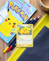 A drawing book featuring Pikachu, a yellow cartoon character, is placed on a wooden surface. Next to it are colored pencils in yellow, blue, and red. There is also a trading card depicting Pikachu, showcasing its abilities. The setup includes a cup of a drink, likely tea or coffee, partially visible at the edge.