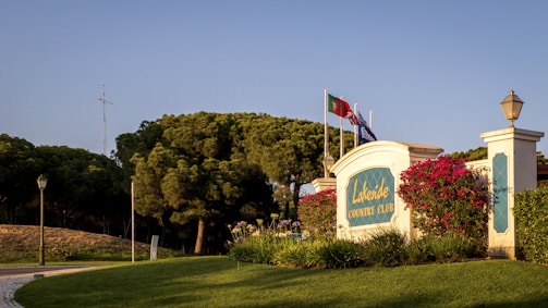 A well-maintained lawn with vibrant green grass surrounds the entrance of a country club. A prominent sign with 'Lakeside Country Club' is visible, flanked by blooming pink flowers and two lantern-style light fixtures. Behind the sign, several flags fly atop white poles. Tall, dense trees form a lush backdrop under a clear blue sky.
