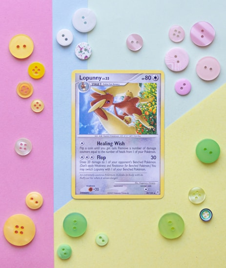 A Pok&eacute;mon trading card, featuring Lopunny, is placed on a background divided into three pastel-colored sections: pink, blue, and yellow. Surrounding the card are various colorful buttons in hues of pink, white, yellow, and green, adding a playful touch.