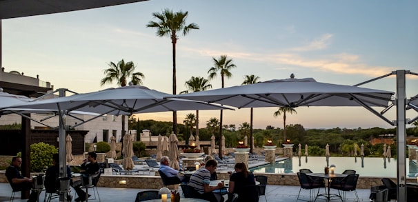 A serene outdoor scene with a hotel pool and palm trees, featuring people seated under large white umbrellas on a patio. The setting sun casts a warm glow on the surroundings, and the pool reflects the sky and adjacent landscape.