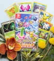 A collection of Pokémon trading cards and a Nintendo Switch game case titled 'New Pokémon Snap.' The cards feature various Pokémon characters surrounded by tropical foliage, colorful flowers, and small ornamental plants.