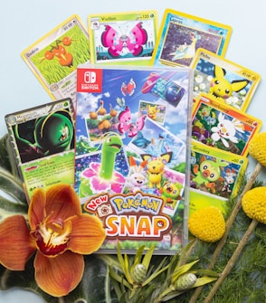 A collection of Pokémon trading cards and a Nintendo Switch game case titled 'New Pokémon Snap.' The cards feature various Pokémon characters surrounded by tropical foliage, colorful flowers, and small ornamental plants.