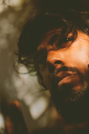 A close-up portrait capturing a thoughtful expression on a man's face. The lighting is soft and dramatic, creating shadows across his features and highlighting his dark hair and beard. The backdrop is blurred, giving a shallow depth of field effect.