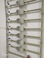 Several water meters are mounted on a wall, connected by a network of pipes. The meters have dials with numerical displays, and there are blue valve handles at the joints. The setup has a uniform arrangement with neat pipe fittings.