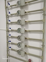 Several water meters are mounted on a wall, connected by a network of pipes. The meters have dials with numerical displays, and there are blue valve handles at the joints. The setup has a uniform arrangement with neat pipe fittings.