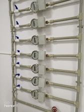 Several water meters are mounted on a wall, connected by a network of pipes. The meters have dials with numerical displays, and there are blue valve handles at the joints. The setup has a uniform arrangement with neat pipe fittings.