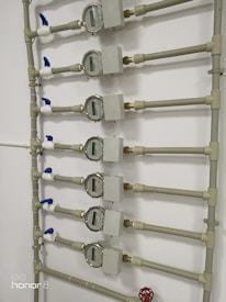 Several water meters are mounted on a wall, connected by a network of pipes. The meters have dials with numerical displays, and there are blue valve handles at the joints. The setup has a uniform arrangement with neat pipe fittings.