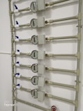 Several water meters are mounted on a wall, connected by a network of pipes. The meters have dials with numerical displays, and there are blue valve handles at the joints. The setup has a uniform arrangement with neat pipe fittings.