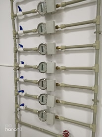 Several water meters are mounted on a wall, connected by a network of pipes. The meters have dials with numerical displays, and there are blue valve handles at the joints. The setup has a uniform arrangement with neat pipe fittings.