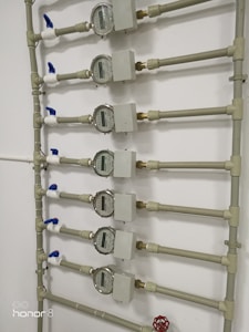 Several water meters are mounted on a wall, connected by a network of pipes. The meters have dials with numerical displays, and there are blue valve handles at the joints. The setup has a uniform arrangement with neat pipe fittings.