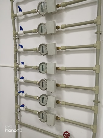 Several water meters are mounted on a wall, connected by a network of pipes. The meters have dials with numerical displays, and there are blue valve handles at the joints. The setup has a uniform arrangement with neat pipe fittings.