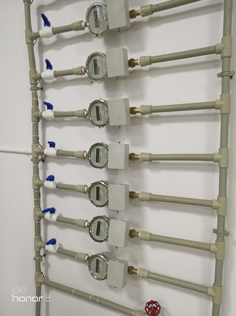 Several water meters are mounted on a wall, connected by a network of pipes. The meters have dials with numerical displays, and there are blue valve handles at the joints. The setup has a uniform arrangement with neat pipe fittings.