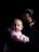 A person wearing a dark face mask is holding an infant wrapped in a pink blanket. The scene is softly lit, highlighting the baby’s face while the background remains dark, creating a sense of intimacy and focus on the subject.