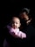 A person wearing a dark face mask is holding an infant wrapped in a pink blanket. The scene is softly lit, highlighting the baby’s face while the background remains dark, creating a sense of intimacy and focus on the subject.
