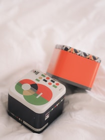 Two decorative tin boxes are resting on a soft, white fabric. One box features a design with geometric shapes and vibrant colors, including green, red, and black, resembling a musical instrument. The other box is primarily orange with a patterned rim.