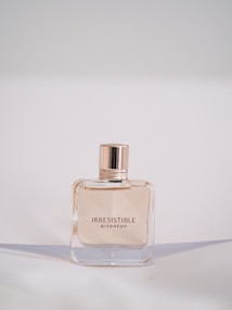 A bottle of Givenchy Irresistible perfume is centered against a soft, neutral background. The bottle has a simple, elegant square design with a glossy, rose-gold cap. The perfume inside is lightly tinted, complementing the sophisticated style.