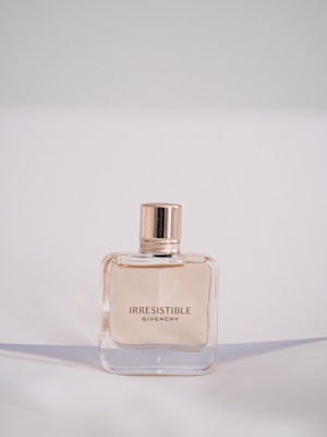 A bottle of Givenchy Irresistible perfume is centered against a soft, neutral background. The bottle has a simple, elegant square design with a glossy, rose-gold cap. The perfume inside is lightly tinted, complementing the sophisticated style.