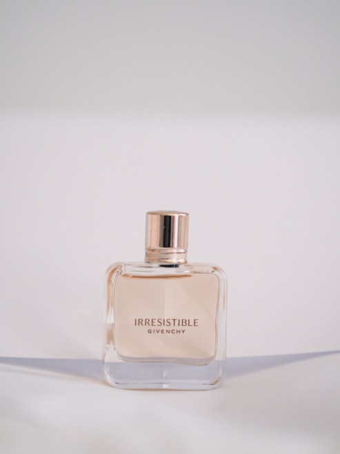 A bottle of Givenchy Irresistible perfume is centered against a soft, neutral background. The bottle has a simple, elegant square design with a glossy, rose-gold cap. The perfume inside is lightly tinted, complementing the sophisticated style.