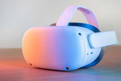 A white VR headset with an adjustable strap is placed on a wooden surface. The device is illuminated by soft, colorful lighting, creating a gradient effect across its surface. The design is sleek and modern, showcasing multiple sensors and a comfortable headband.