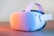 A white VR headset with an adjustable strap is placed on a wooden surface. The device is illuminated by soft, colorful lighting, creating a gradient effect across its surface. The design is sleek and modern, showcasing multiple sensors and a comfortable headband.