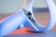 A close-up of a modern gaming controller, primarily in light blue with some buttons and a circular handle. The lighting casts a soft pink and blue glow, highlighting its sleek design.
