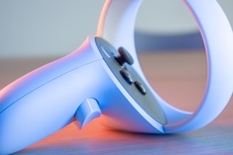 A close-up of a modern gaming controller, primarily in light blue with some buttons and a circular handle. The lighting casts a soft pink and blue glow, highlighting its sleek design.